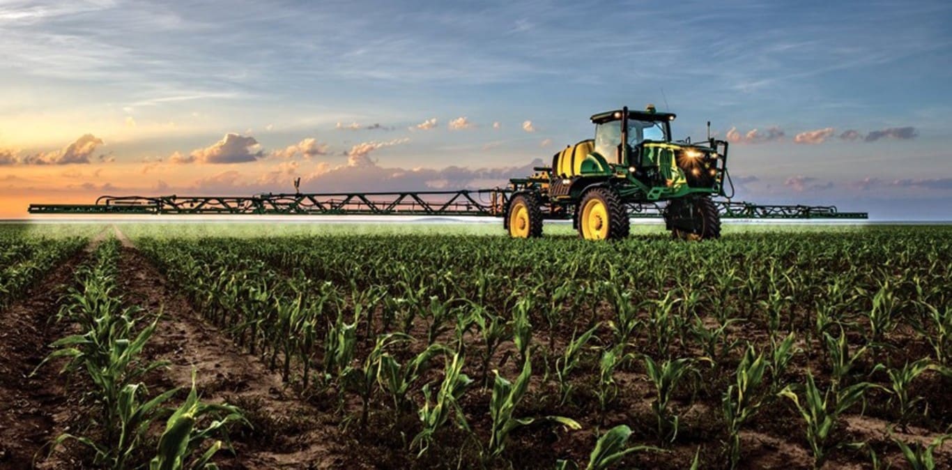 John Deere sproeier, werkend in een maïsveld met de zonsondergang op de achtergrond