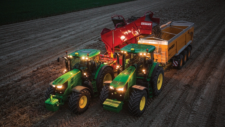 Tractor die 's nachts een aardappeloogstmachine trekt is aardappelen aan het lossen naar een wagen die door een andere tractor wordt getrokken