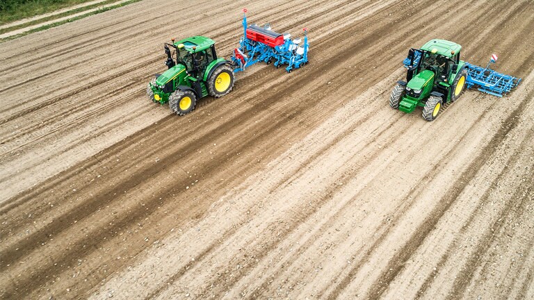 Twee John Deere 6M-tractoren op een braakliggend veld De ene trekt een Monsomen-zaaimachine. De andere trekt een Lemken-zaaibedcombinatie