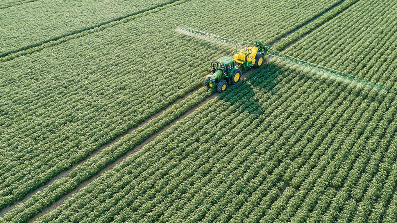 Een John Deere 6M-reeks tractor die een M700-veldspuit trekt door een aardappelveld.