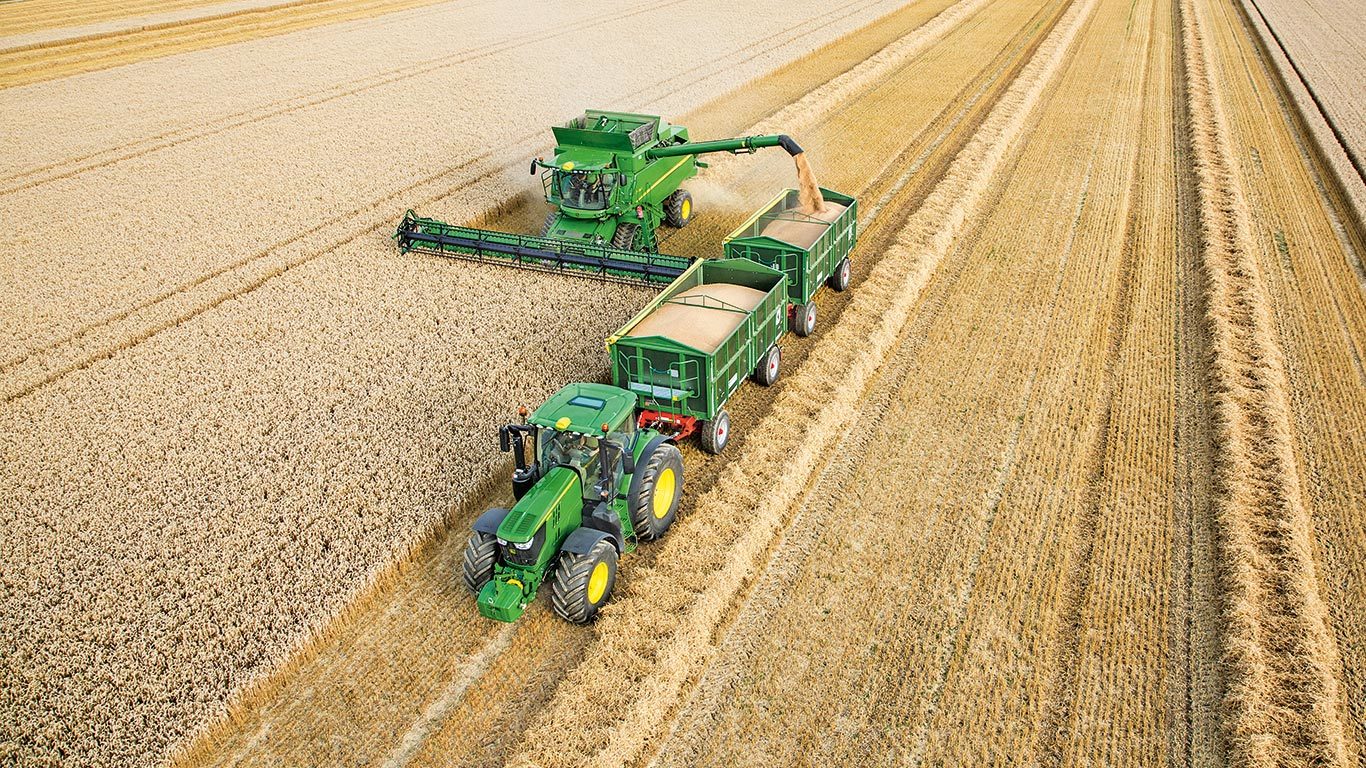 John Deere S-Series maaidorser die tarwe lost in dubbele aanhangwagens getrokken door een tractor