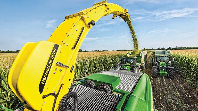 Zicht vanaf de achterkant van John Deere 9000 Series zelfrijdende veldhakselaar met twee kuilvoerwagens getrokken door John Deere tractors direct erachter