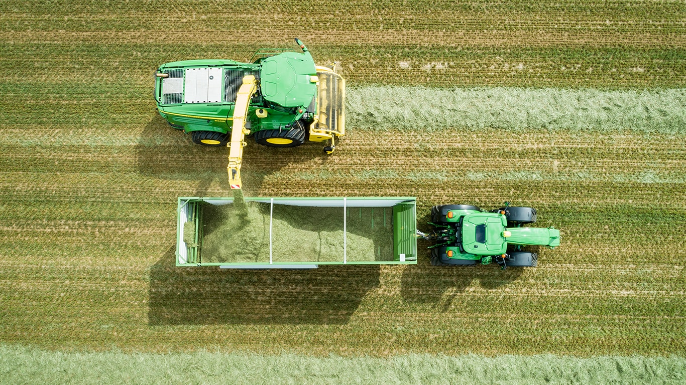 Luchtfoto van John Deere 8000 Series zelfrijdende veldhakselaar met een gras pick-up die kuilvoer laadt in een wagen getrokken door een John Deere tractor