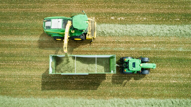 Luchtfoto van John Deere 8000 Series zelfrijdende veldhakselaar met een gras pick-up die kuilvoer laadt in een wagen getrokken door een John Deere tractor