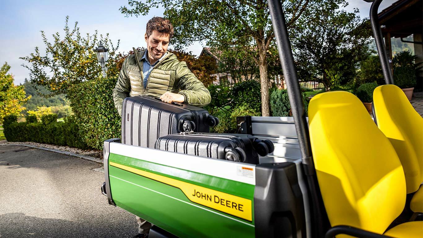 Persoon die een grote zwarte koffer laadt in de laadbak van een John Deere Gator GS met felgele stoelen, omringd door groen en bomen.
