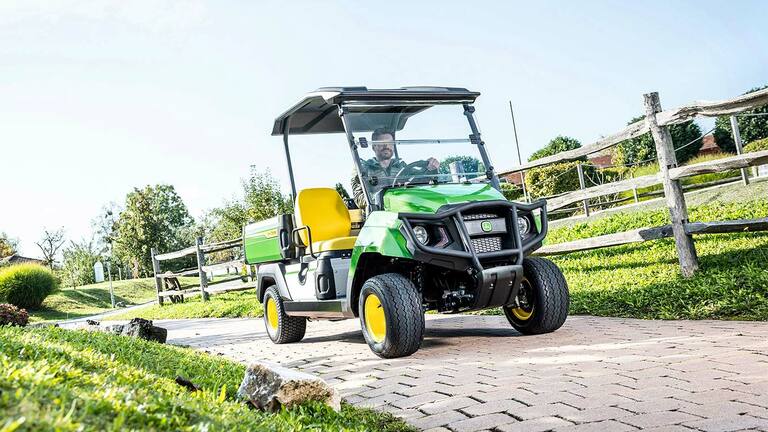 Groen universeel John Deere Gator GS-voertuig die rijdt op een verhard pad door een aangelegd gebied met gras en houten hekken.