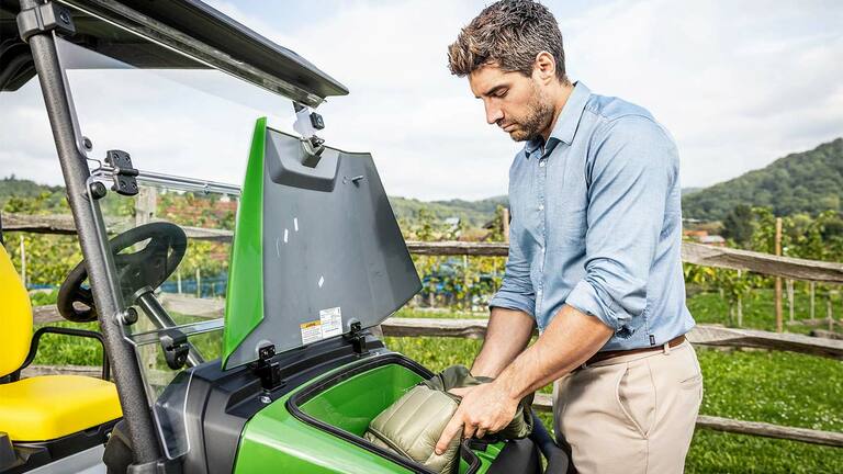 Persoon die een gevoerde tas plaatst in het voorste opbergvak van een groene John Deere Gator GS die buiten geparkeerd staat, vlakbij een houten hek en een wijngaard.