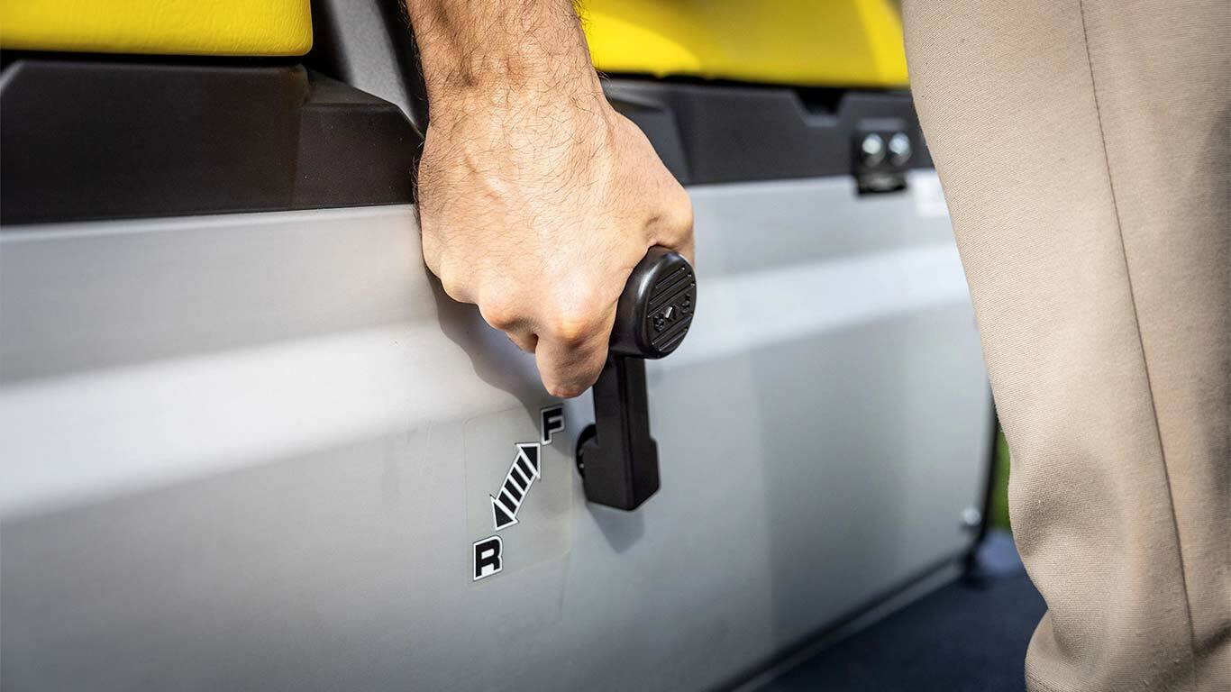 Close-up van een hand die de versnellingshendel bedient op een John Deere Gator GS, met de posities vooruit en achteruit naast gele stoelen.