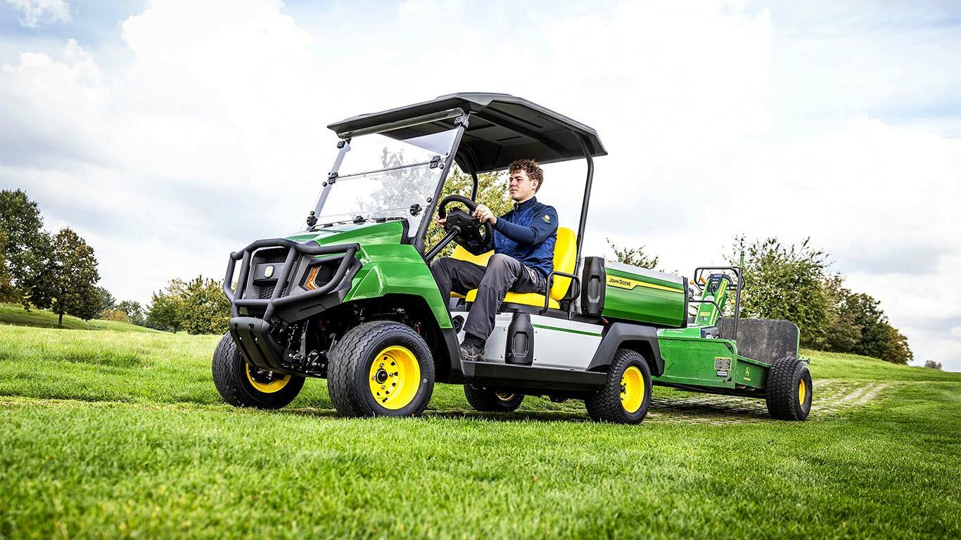 Groen John Deere Gator elektrisch universeel voertuig die apparatuur sleept over een grasveld onder een deels bewolkte lucht.