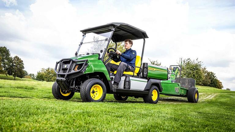 Groen John Deere Gator elektrisch universeel voertuig die apparatuur sleept over een grasveld onder een deels bewolkte lucht.