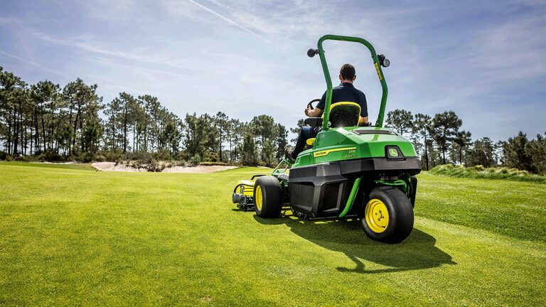 Een persoon rijdt met een groene John Deere 2775 E-Cut™ Electric greens maaier over een goed onderhouden golfbaan, met bomen en een deels bewolkte lucht op de achtergrond.