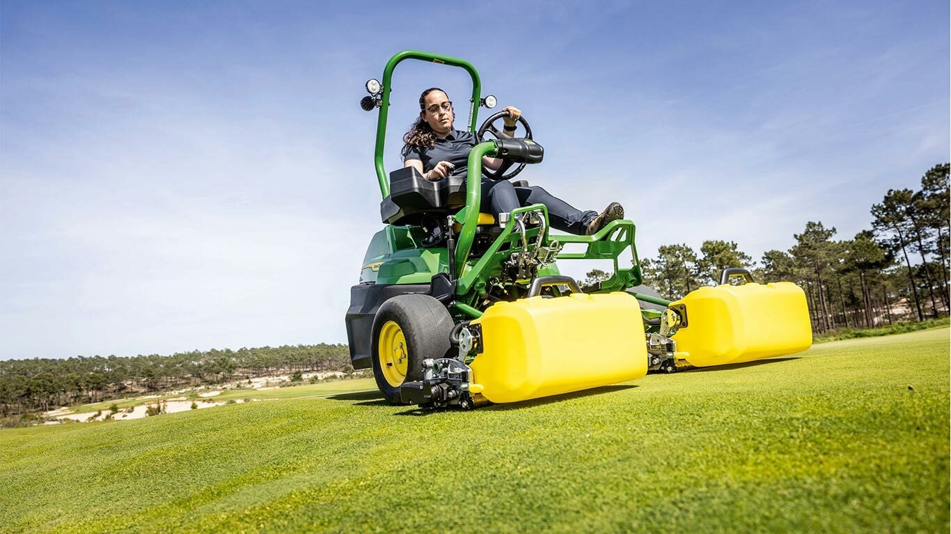 Een persoon bedient een groene en gele John Deere 2775 E-Cut™ Electric maaier met voorrollen bedekt met een gele behuizing op een golf green, met bomen en een heldere blauwe lucht op de achtergrond.