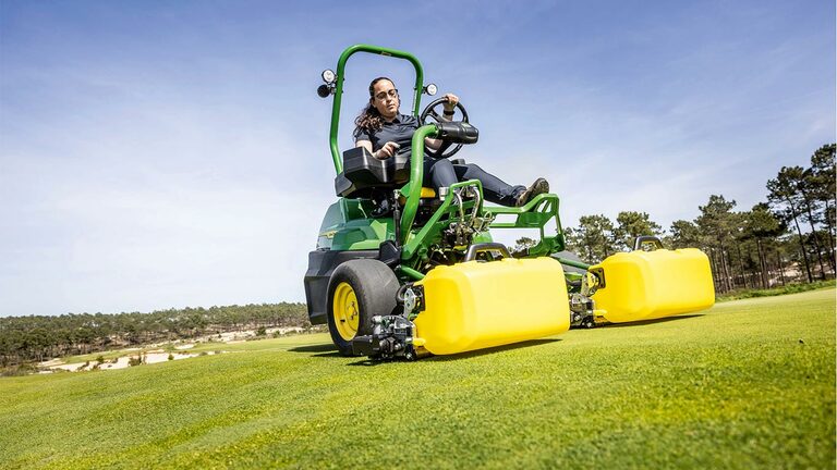 Een persoon bedient een groene en gele John Deere 2775 E-Cut™ Electric maaier met voorrollen bedekt met een gele behuizing op een golf green, met bomen en een heldere blauwe lucht op de achtergrond.