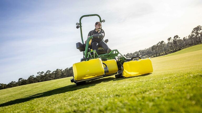 Een bestuurder werkt met een groen-gele John Deere 2775 E-Cut™ Electric maaier, uitgerust met drie grote voorrollen die zijn afgeschermd met gele behuizingen, op een golfgreen, met bomen en een deels bewolkte lucht op de achtergrond.