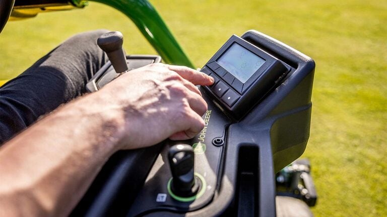 Close-up van de hand van een persoon met het TechControl-paneel op een John Deere 2775 E-Cut™ Electric maaier, drukt op knoppen op een digitaal display met een joystick en andere bedieningselementen zichtbaar.