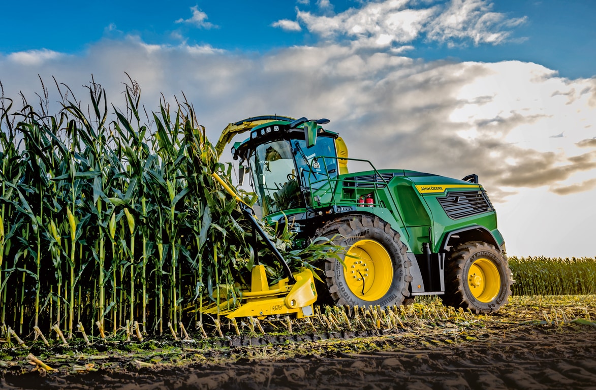 Een veldhakselaar van John Deere oogst mais in een veld met daarboven een blauwe lucht met wolken.