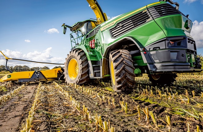Een John Deere veldhakselaar aan het werk in een geoogst maisveld, waarbij de grote banden en de snij-inrichting van de machine worden getoond tegen een heldere blauwe lucht.