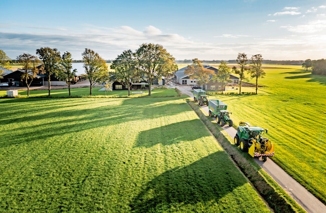 Een luchtfoto van een boerderij met groene velden, bomen en meerdere tractoren en zelfrijdende veldhakselaars op een smalle weg die leidt naar een boerderij onder een heldere lucht.