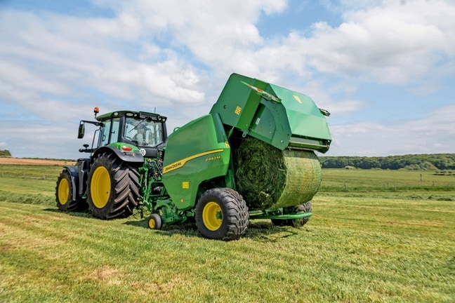 John Deere F442M-balenpers getrokken door een John Deere 6M-tractor die een grasbaal uitwerpt