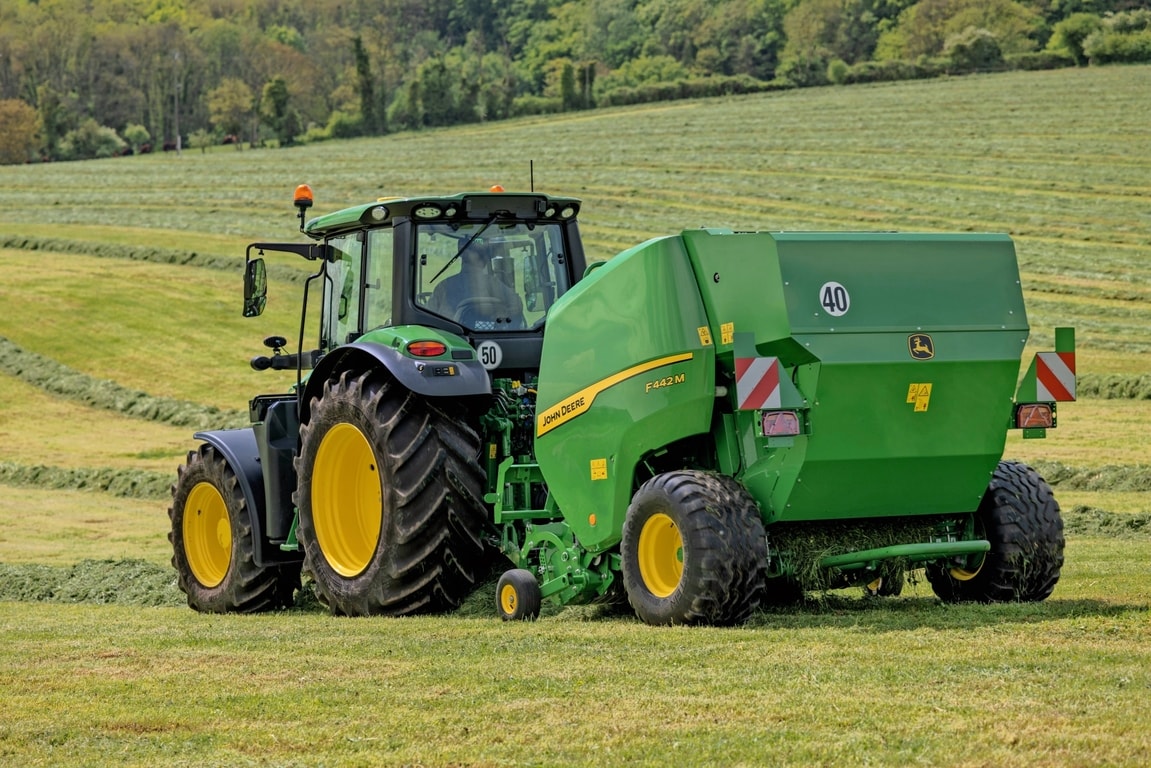 John Deere F442M-balenpers getrokken door een John Deere 6M-tractor op een heuvelachtig grasveld