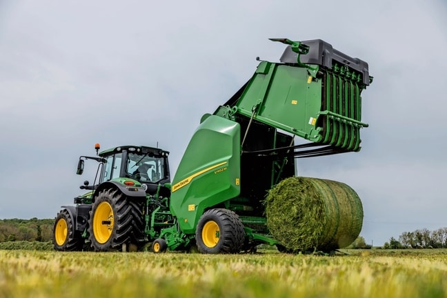 John Deere V462M-balenpers getrokken door een John Deere 6M-tractor die een grasbaal uitwerpt